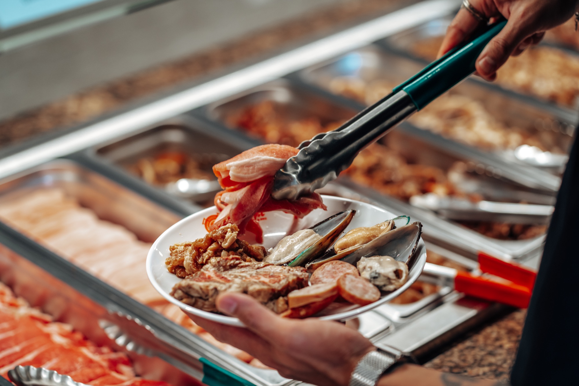 A diner using tongs to load fresh sliced beef, mussels and sausage onto a plate at Lanjie's cold bar